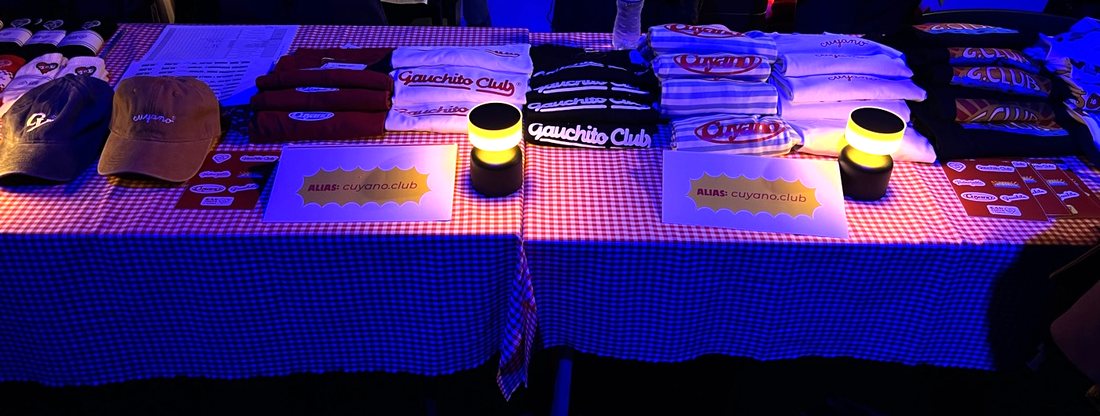 Dos mesas juntas con mantel cuadrillé, pilas de remeras por modelo, gorras, calcos y dos carteles con el alias